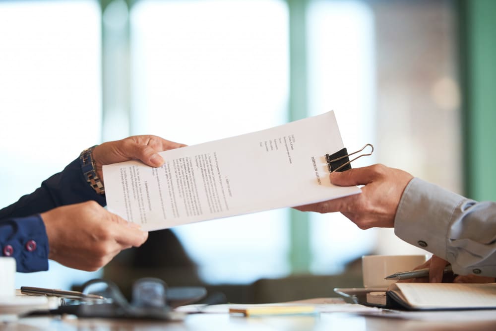 Two people exchanging a contract document with a binder clip in an office setting.