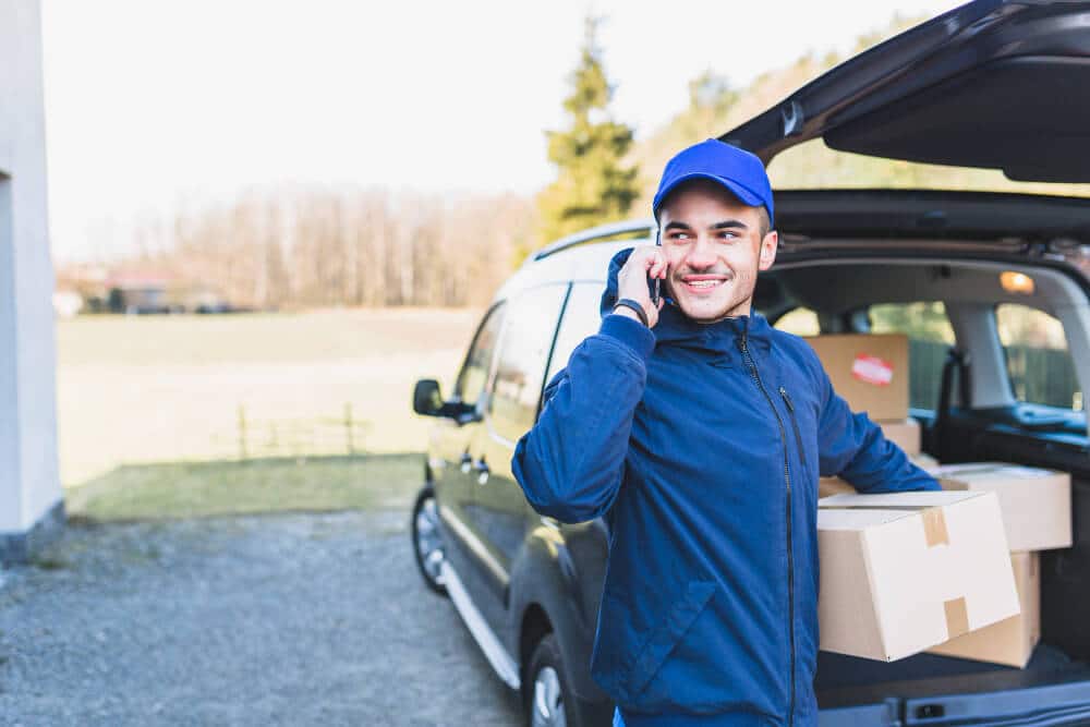 Courier driver holding packages and coordinating route via phone next to delivery van