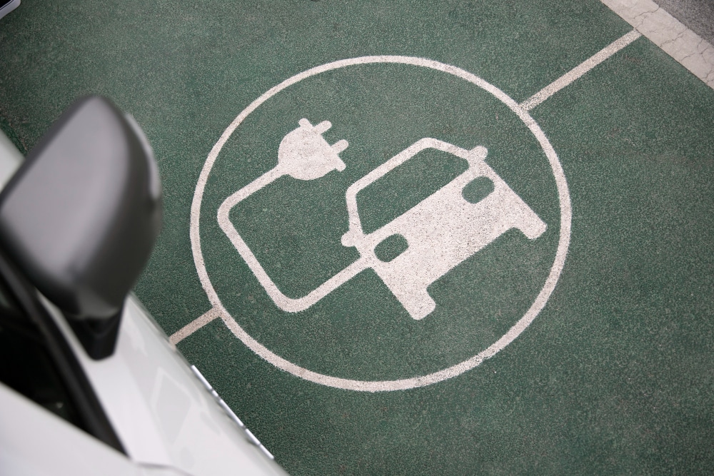 Electric vehicle charging station symbol painted on a green parking space, partially visible next to a white car.