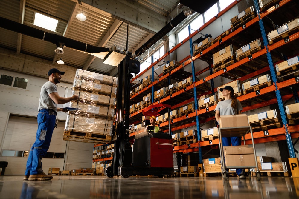 Workers moving pallets and boxes inside a warehouse with tall storage racks.