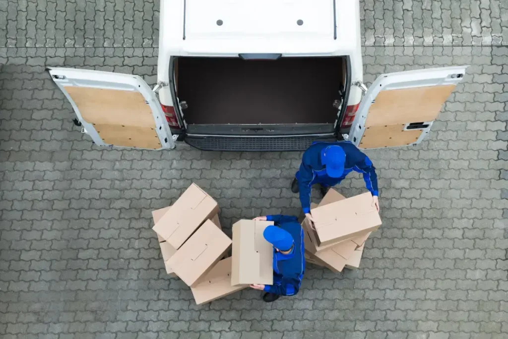 Two delivery workers in blue uniforms load cardboard boxes into a white van during last-mile delivery.