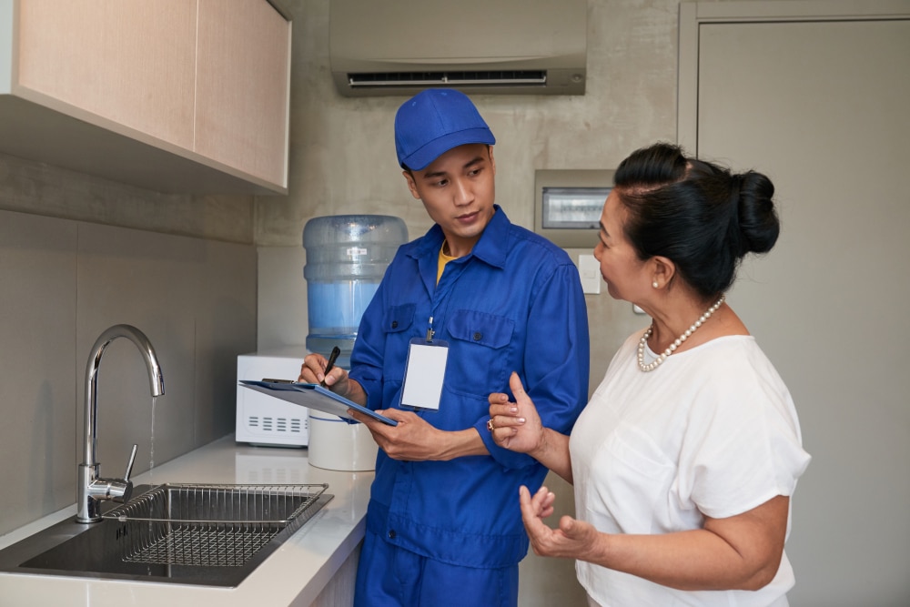Plumber in uniform speaking with a homeowner during a scheduled service visit inside a kitchen.